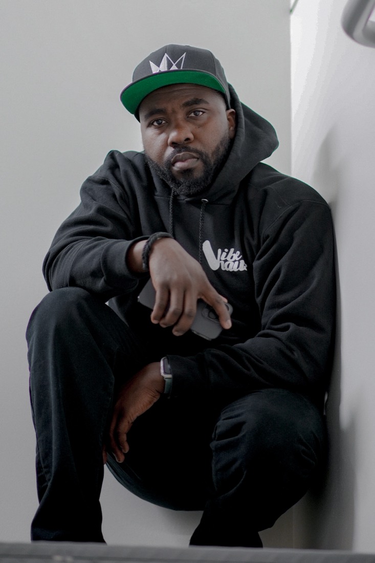 DJ OMG PRINCE crouching in a Vibehauss hoodie and green-brim cap